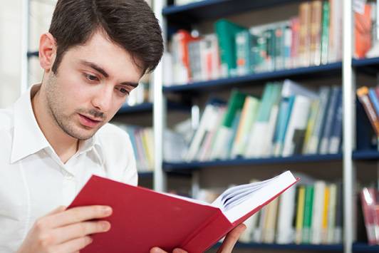 Interessiert lesender junger Mann vor einem Bücherregal.