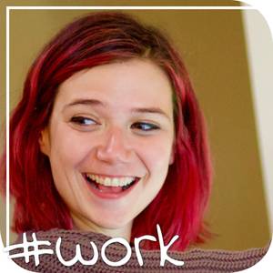 Junge Frau mit roten Haaren steht vor einer Wand mit Klebezetteln und lächelt in die Kamera