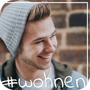 Junger Mann mit Mütze steht vor eine Backsteinwand und lacht.